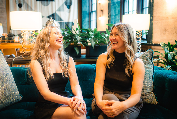 Two women laughing together on a teal sofa in a hotel lounge
